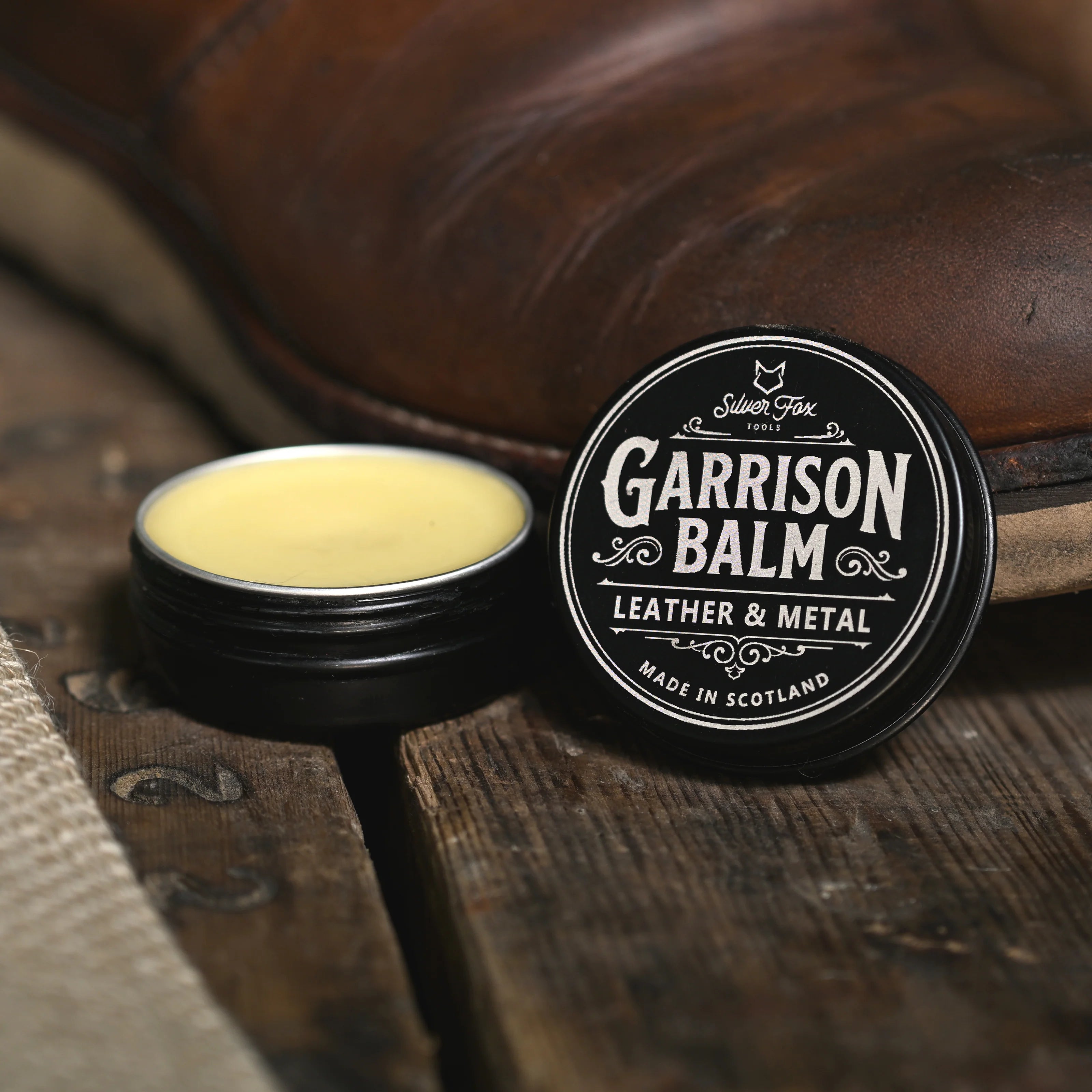 An open tin of Garrison Balm leather and metal protector with the lid standing up by the side leaning on an old leather boot.